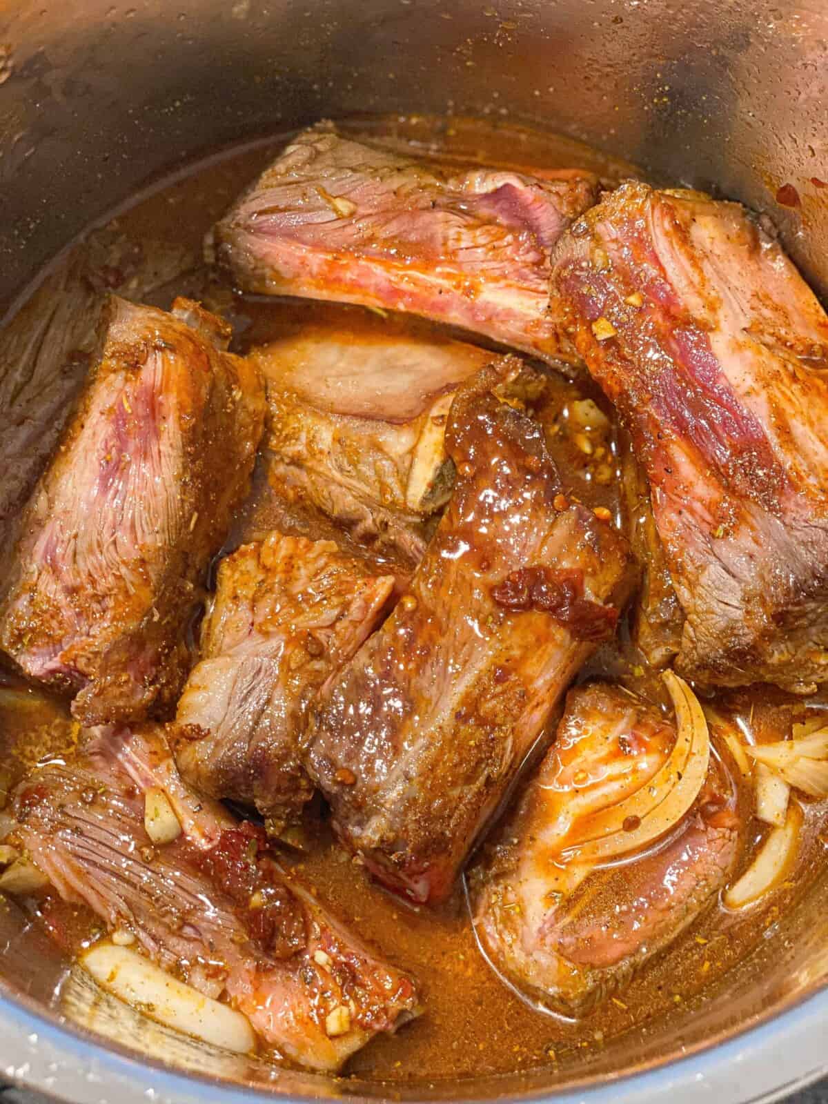 Overhead image of ingredients for Instant Pot short rib barbacoa, including bone-in beef short ribs, sliced onion, garlic, chipotle peppers in adobo, lime, beef broth, honey, soy sauce, Worcestershire sauce, and warm spices.