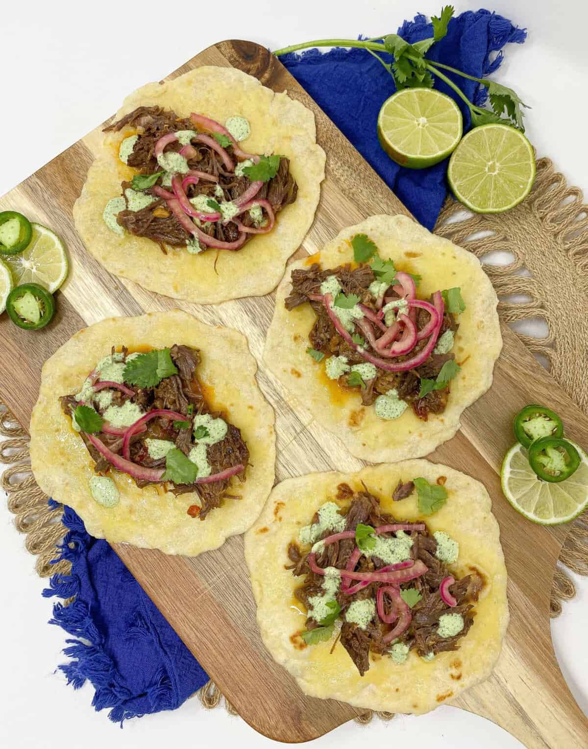 Overhead image of short rib barbacoa tacos topped with shredded beef, fresh cilantro, sliced onion, and lime wedges on warm tortillas.