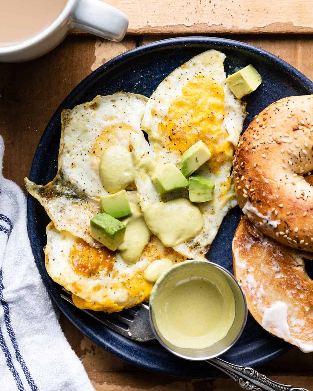 overhead shot fried eggs over hard with avocado, hot sauce, and a bagel