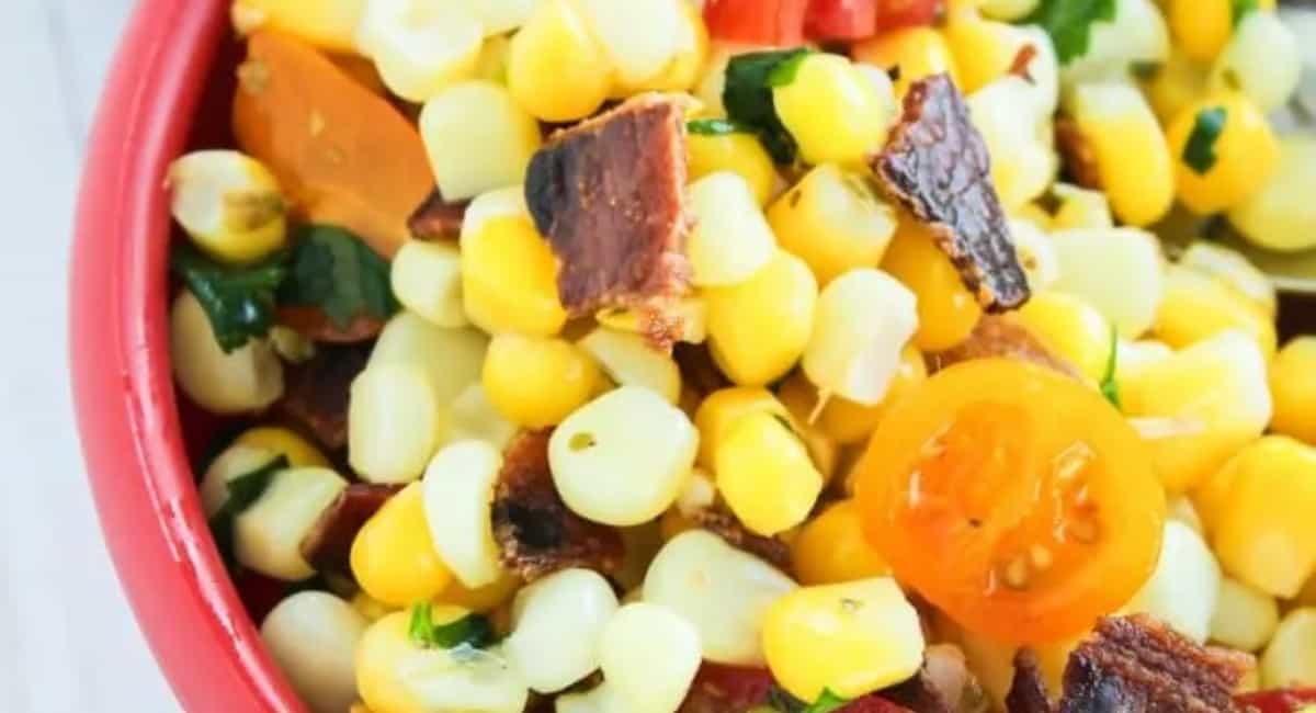 Corn salad topped with bacon and tomatoes and served in a red bowl.