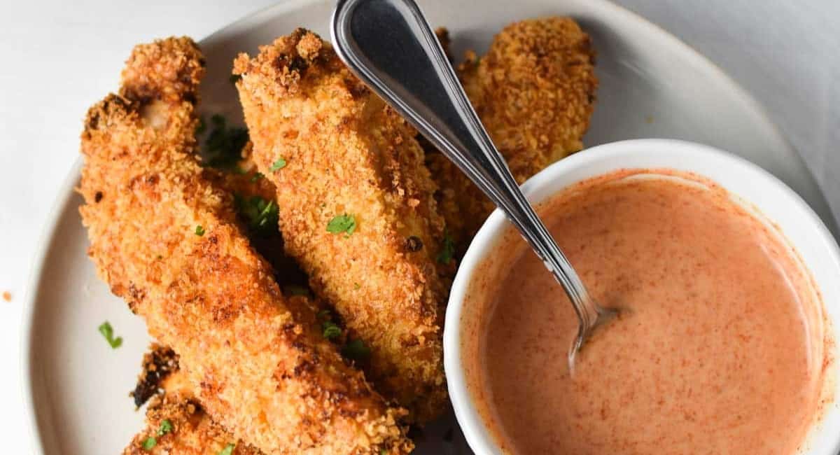 Crispy chicken tenders served on a white plate and with a homemade honey mustard sauce in a bowl.