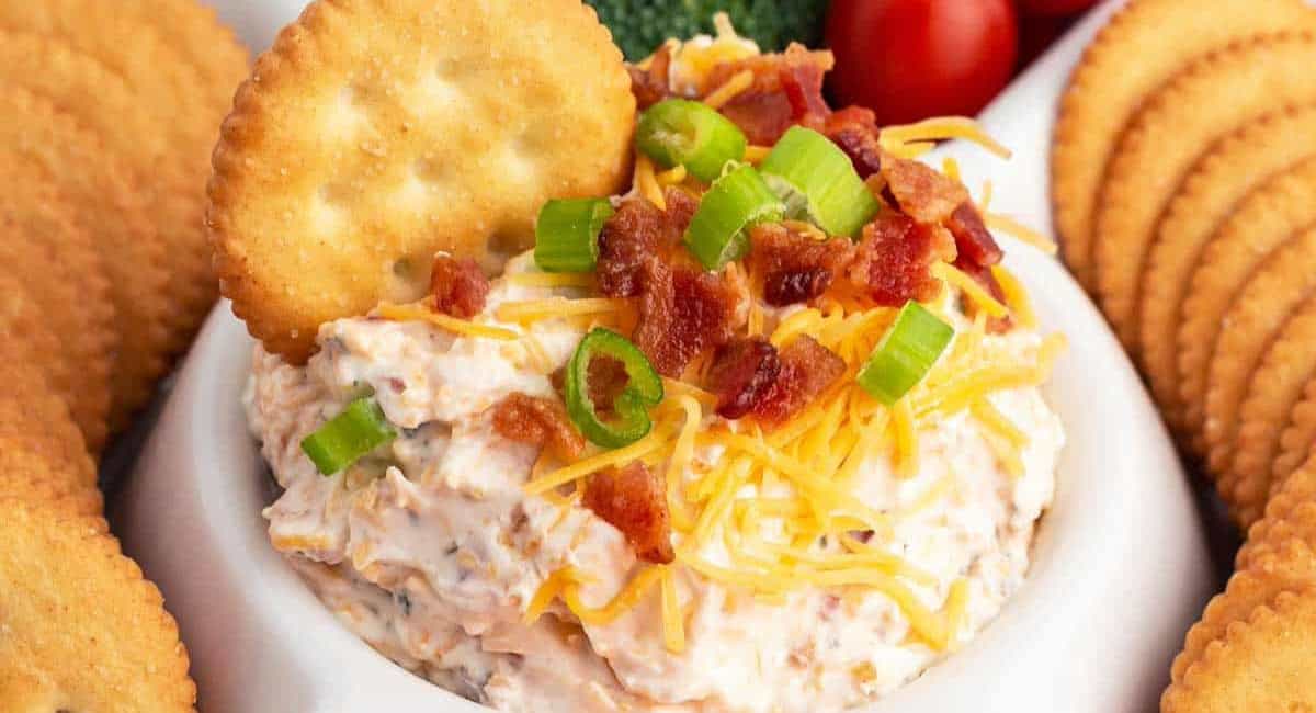 Up close image of bacon cheddar ranch dip with garnish and crackers around small white bowl. 