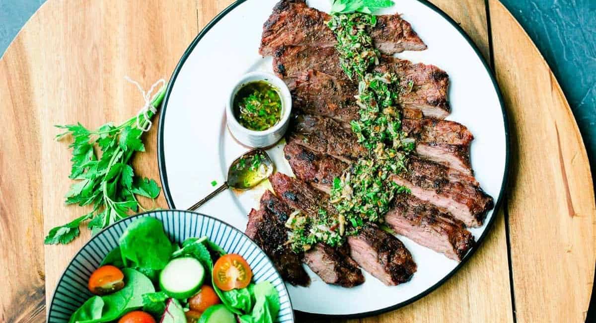 Overhead image of skirt steak with chimichurri sauce on white plate with salad as a side dish. 