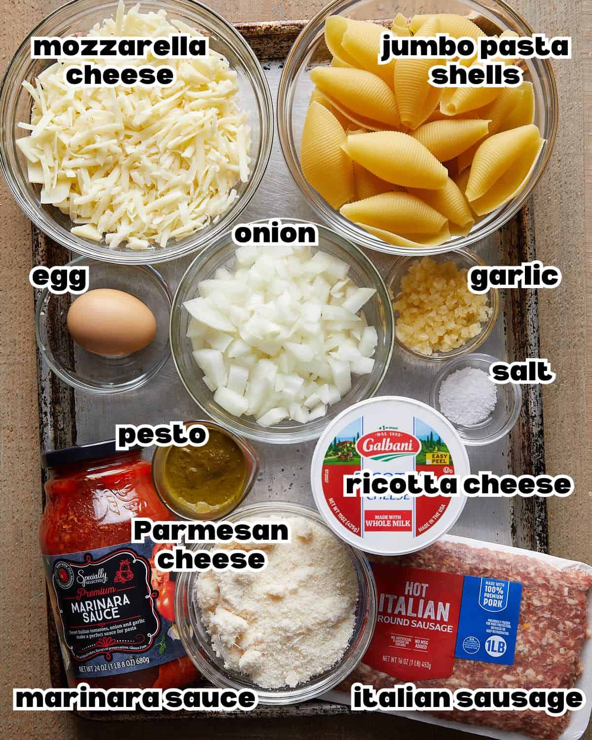 Ingredients needed to make baked stuffed shells, including jumbo pasta shells, ricotta cheese, mozzarella, marinara sauce, eggs, garlic, and pesto.