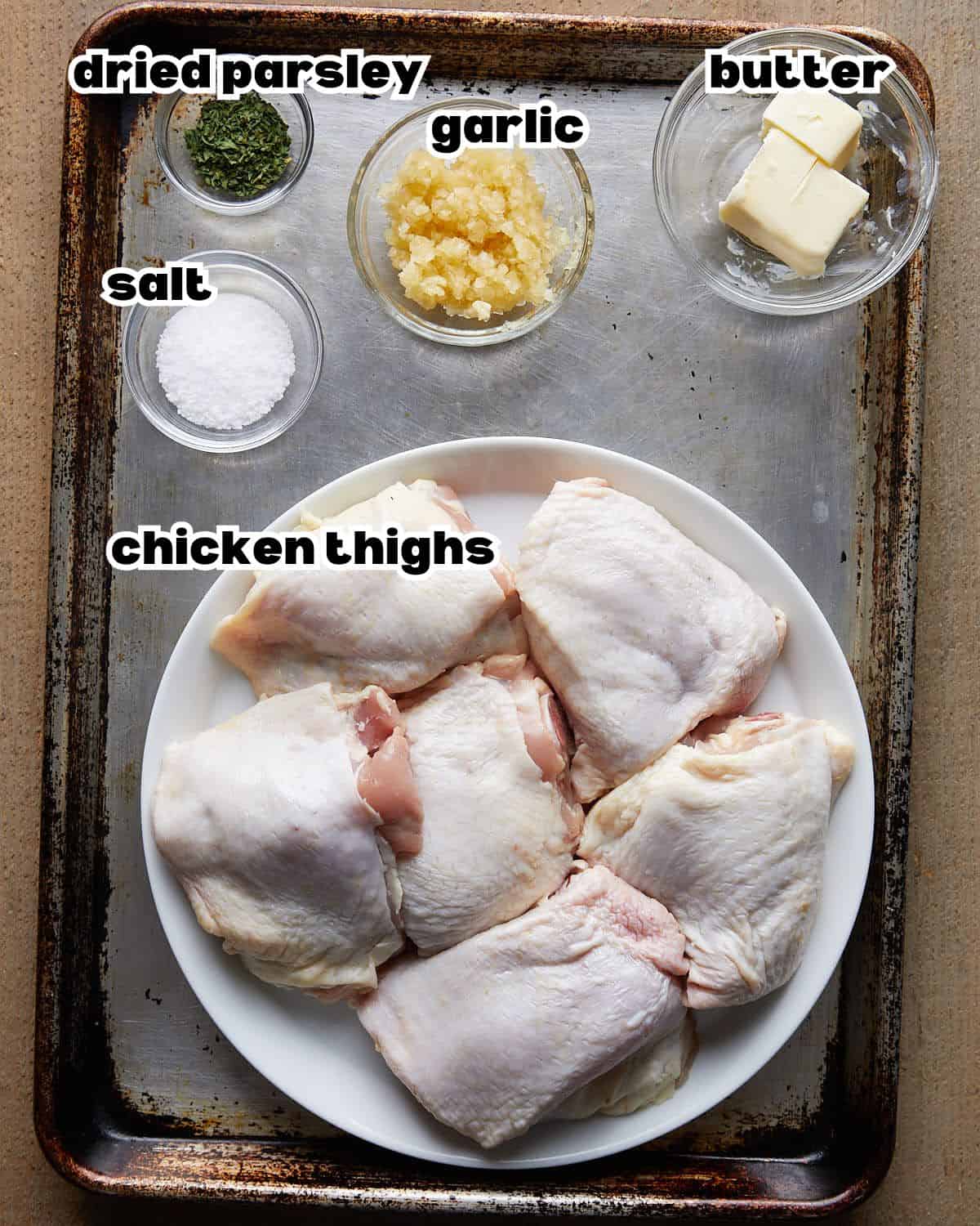 Overhead shot of ingredients for garlic butter chicken thighs, including raw chicken thighs, butter, fresh garlic, and seasonings arranged on a baking sheet.