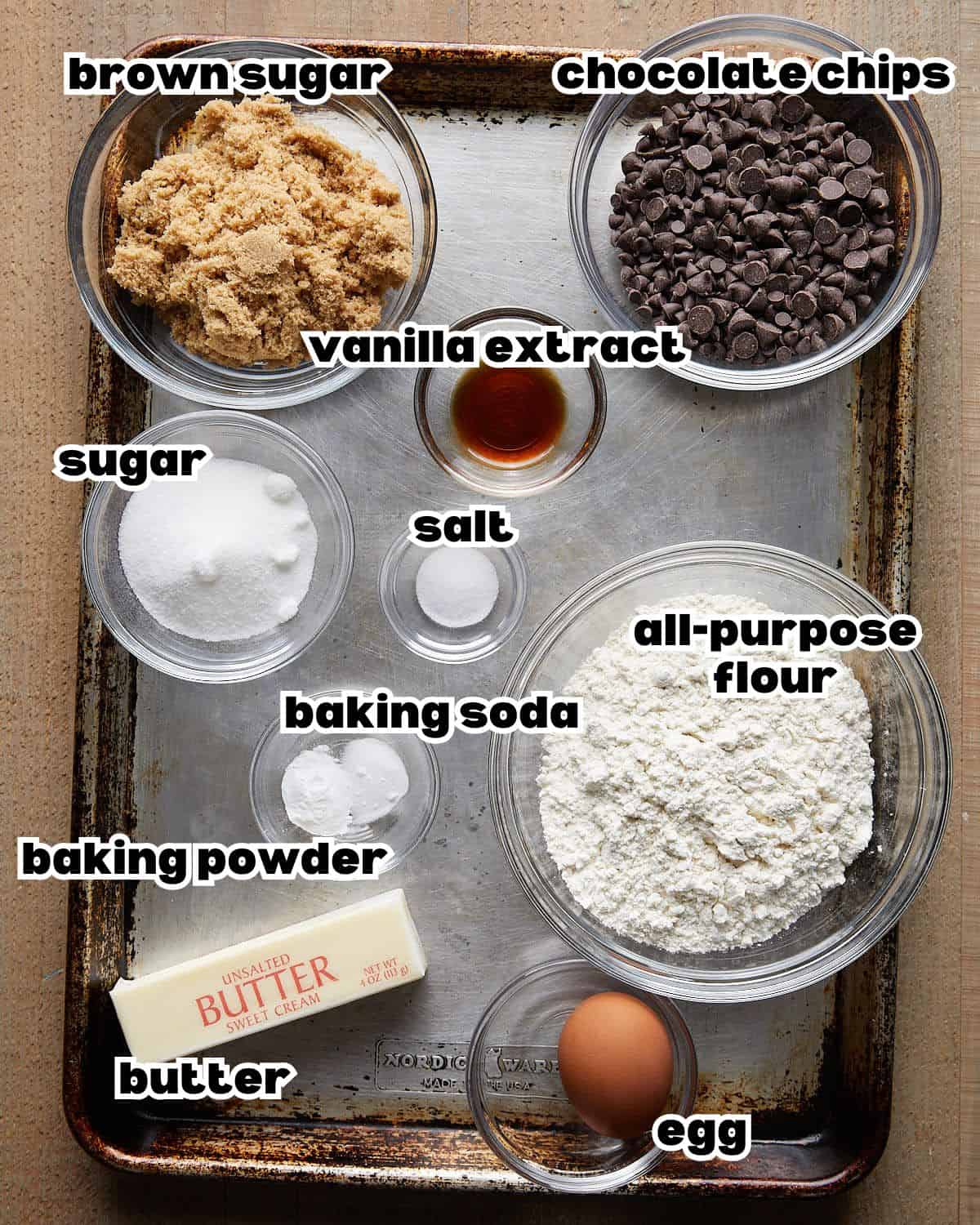 Overhead image of ingredients needed to make no chill chocolate chip cookies, including flour, butter, sugar, eggs, vanilla extract, baking soda, salt, and chocolate chips arranged on a baking sheet.