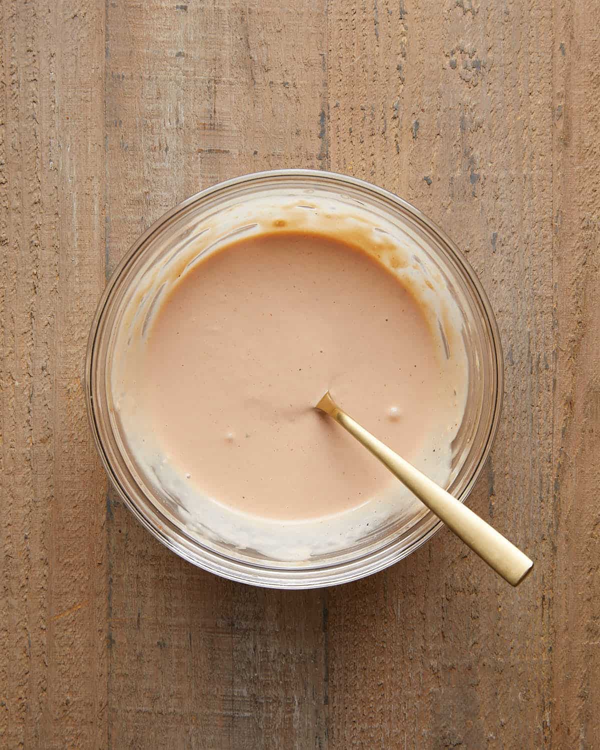 Glass bowl of creamy burger sauce mixed with a gold spoon on a wooden surface.
