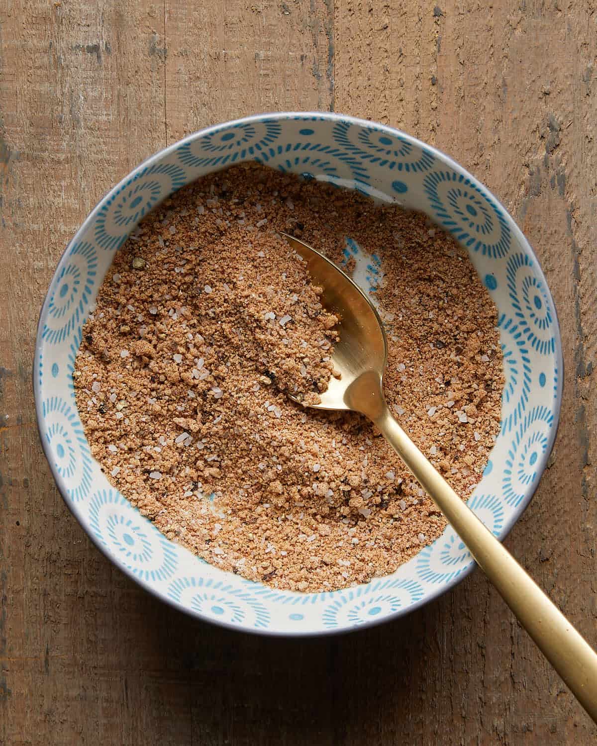 Overhead photo of a gold metal spoon resting in a bowl of burger seasoning on a light background.