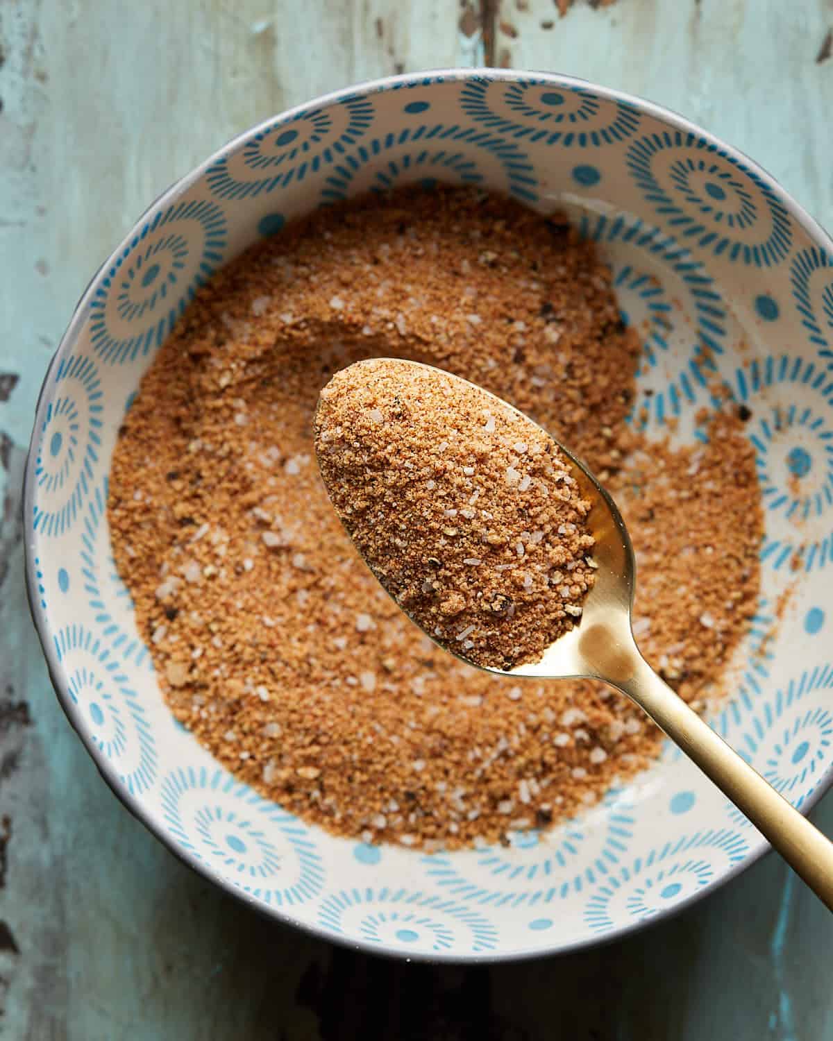 Overhead photo of a bowl of burger seasoning with a gold metal spoon on a rustic surface.