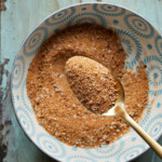 Overhead photo of a bowl of burger seasoning with a gold metal spoon on a rustic surface.