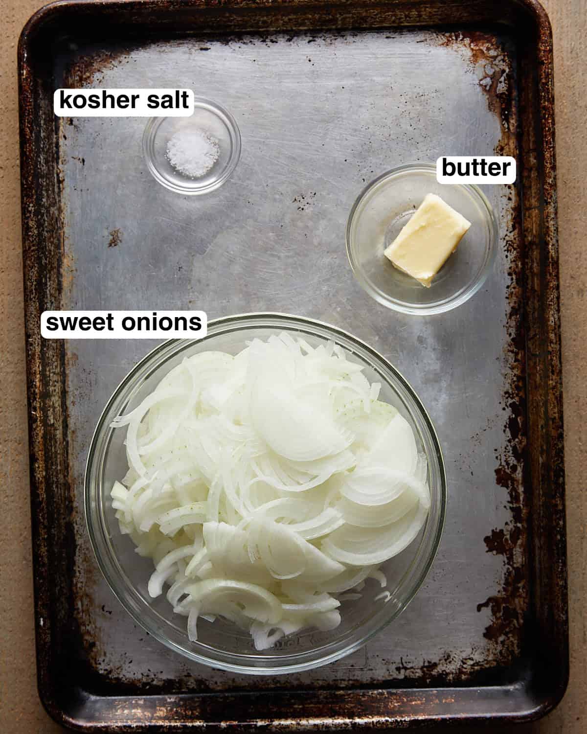 Labeled ingredients for caramelized onions arranged on a sheet pan, including sliced onions, butter, and kosher salt.