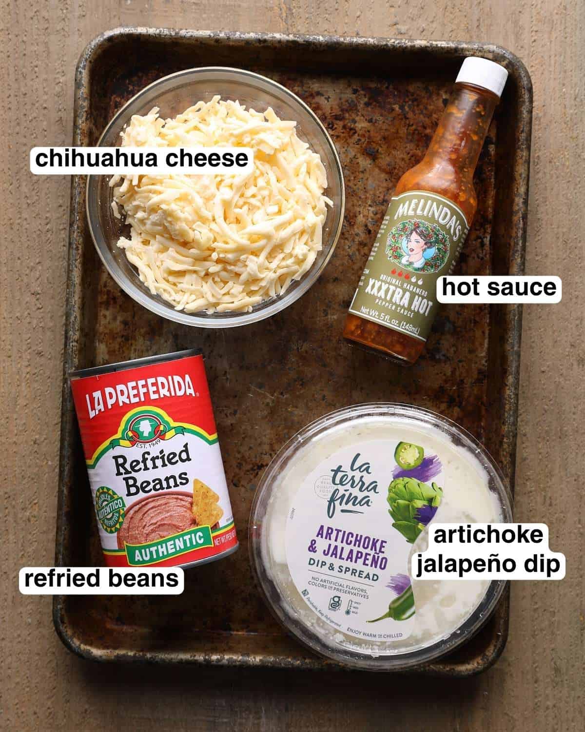 Labeled ingredients for cheesy bean dip, including refried beans, jalapeño artichoke dip, Chihuahua cheese, and hot sauce.