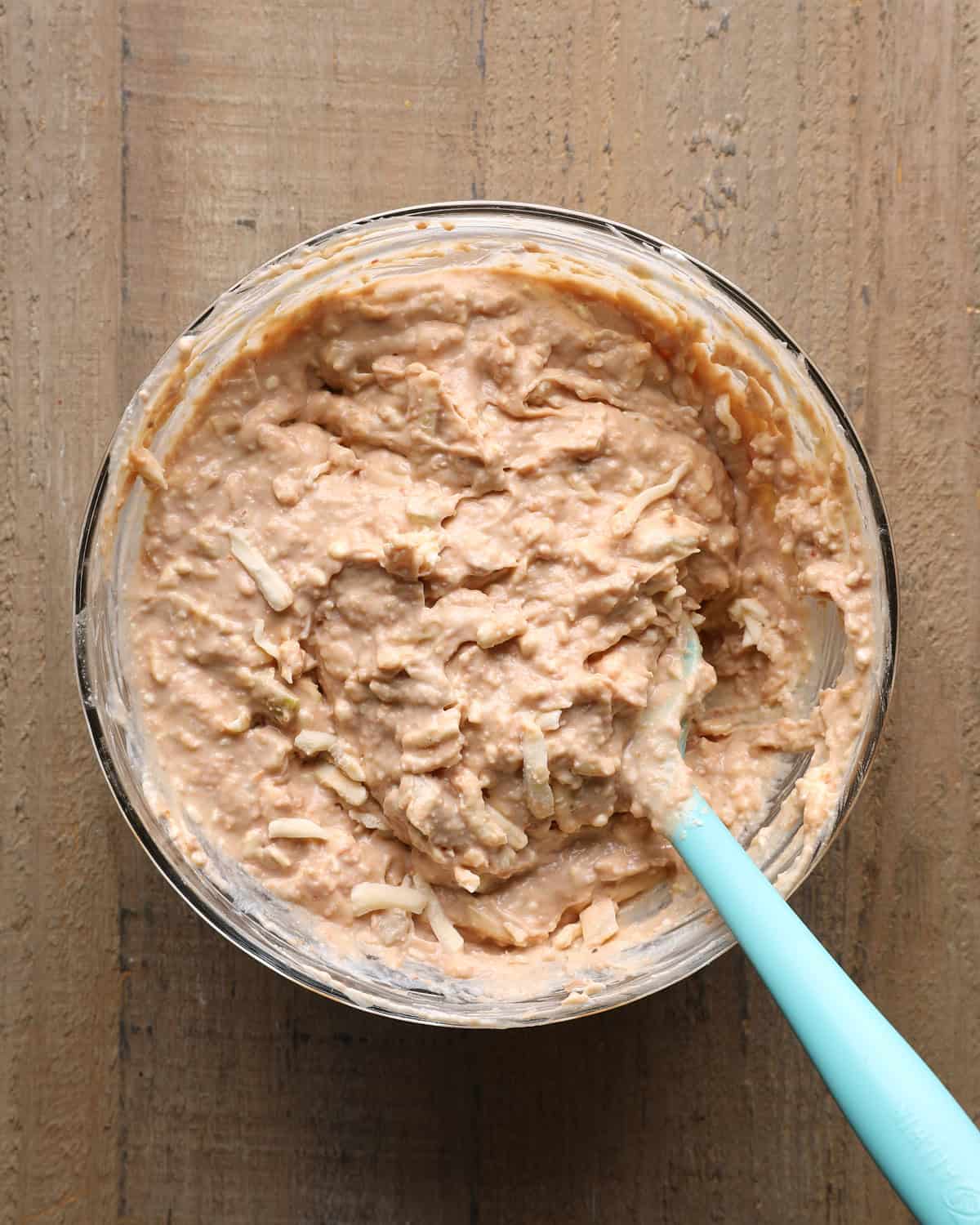 Mixed cheesy bean dip in a glass bowl with a blue spatula before being transferred to a baking dish.