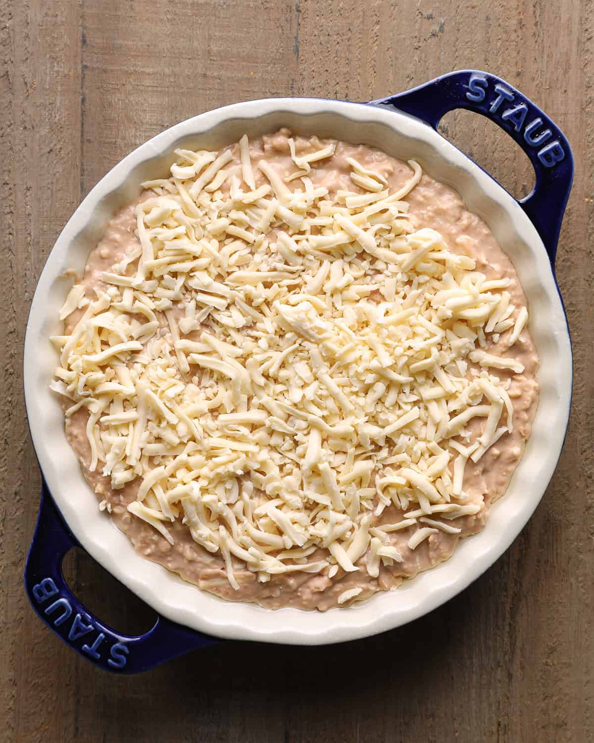 Unbaked cheesy bean dip topped with shredded Chihuahua cheese in a white pie dish.