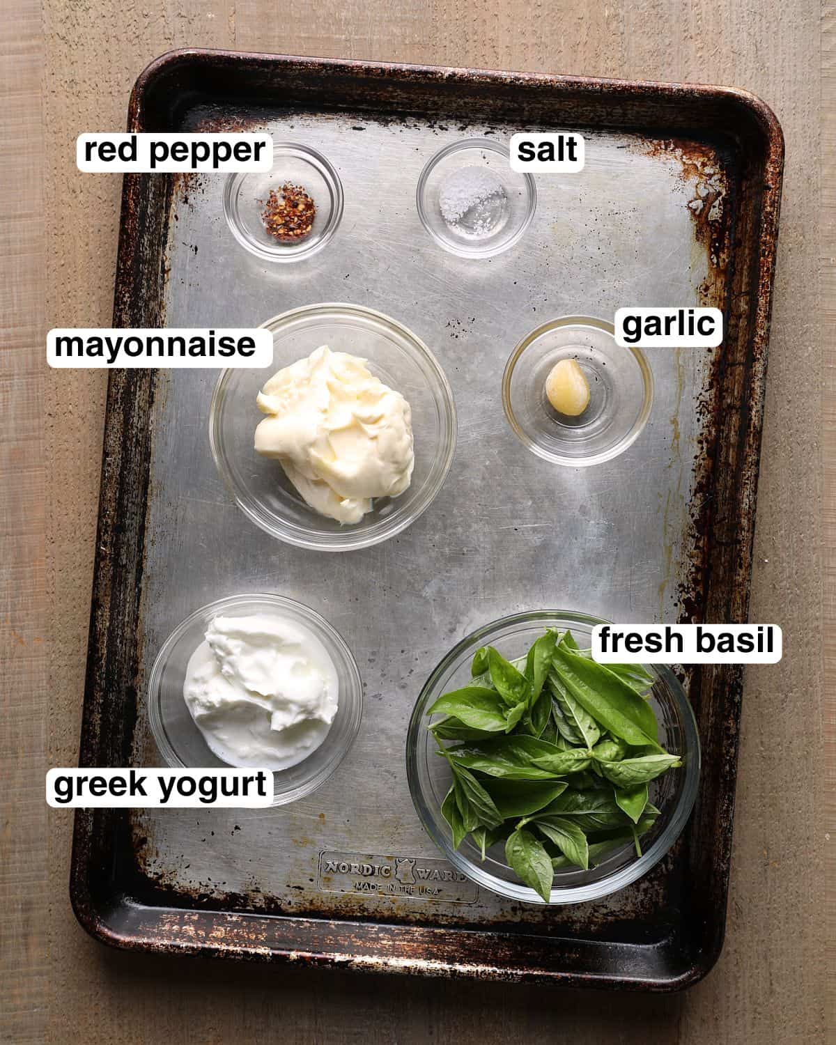 Labeled ingredients for basil aioli arranged on a sheet pan, including mayonnaise, Greek yogurt, fresh basil, garlic, salt, and red pepper flakes.