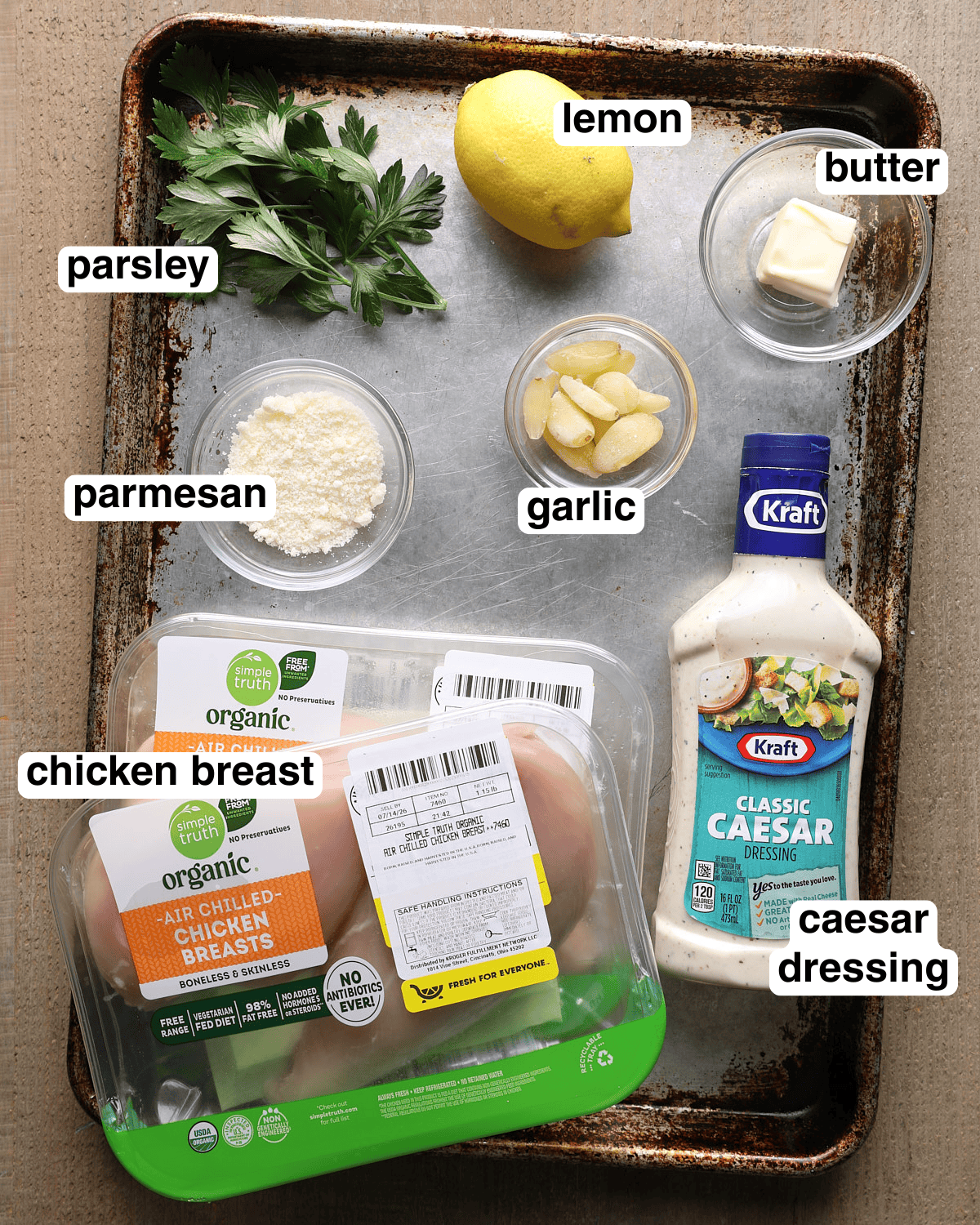 Labeled ingredients for Caesar grilled chicken arranged on a sheet pan, including chicken breast, Caesar dressing, garlic, lemon, parmesan, parsley, and butter.