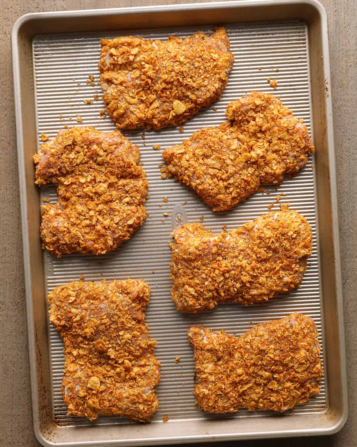 Six cornflake-coated chicken thighs arranged on a baking sheet before baking.