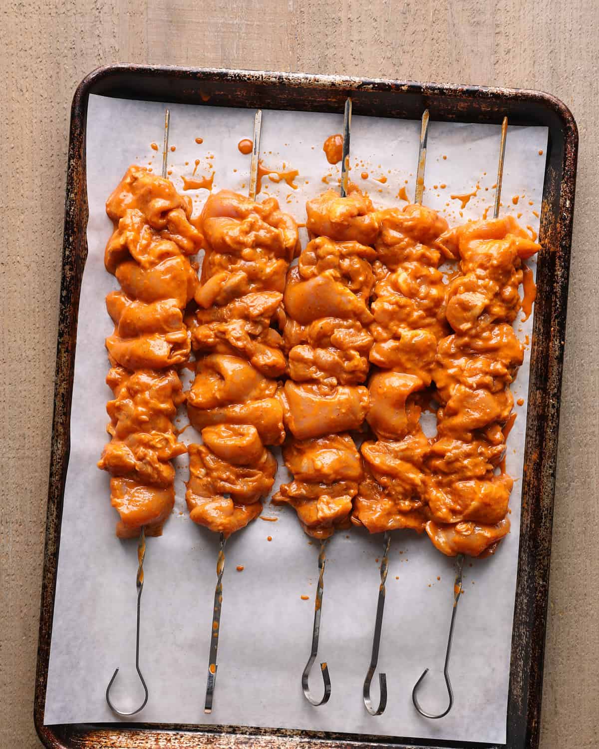 Marinated chicken thigh pieces threaded onto metal skewers on a sheet pan before grilling.