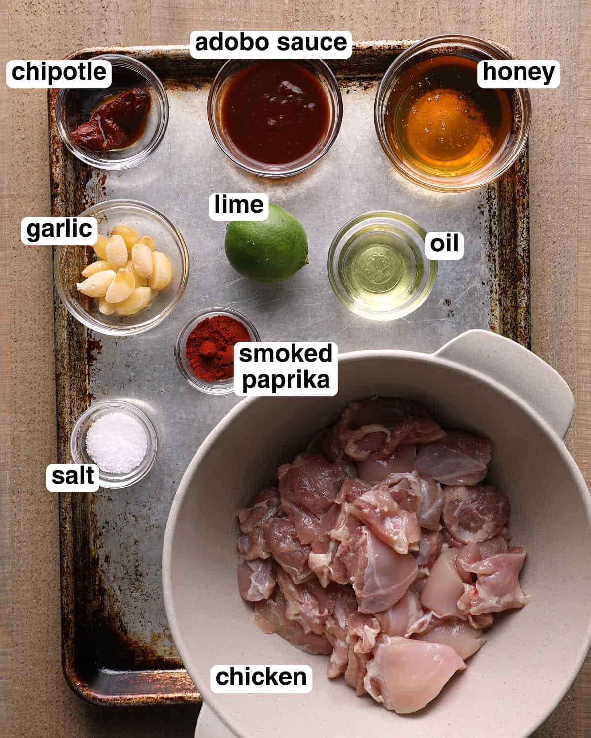 Labeled ingredients for chipotle lime chicken skewers arranged on a sheet pan, including chicken thighs, chipotle peppers, honey, lime, oil, smoked paprika, salt, and garlic.