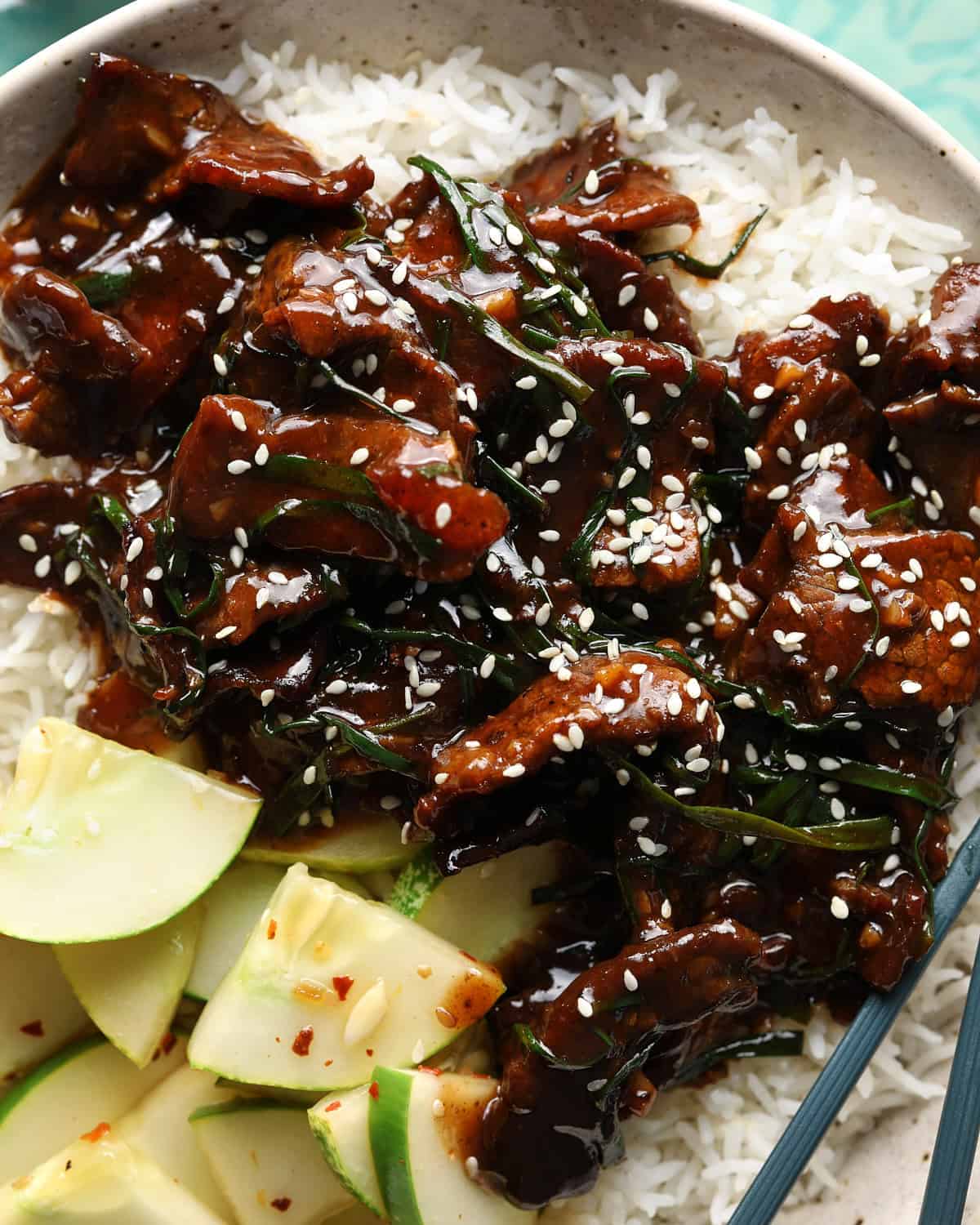 Close-up of Mongolian beef served over rice with cucumbers, green onions, and sesame seeds.