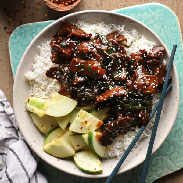 Mongolian beef served over rice with cucumber slices, green onions, and sesame seeds.