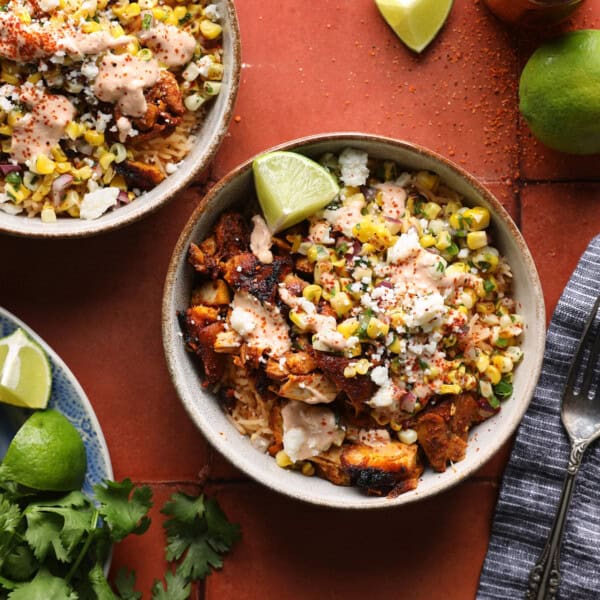 Cropped overhead photo of a street corn chicken rice bowl with grilled chicken, corn salsa, crema, Cotija cheese, and lime wedges.