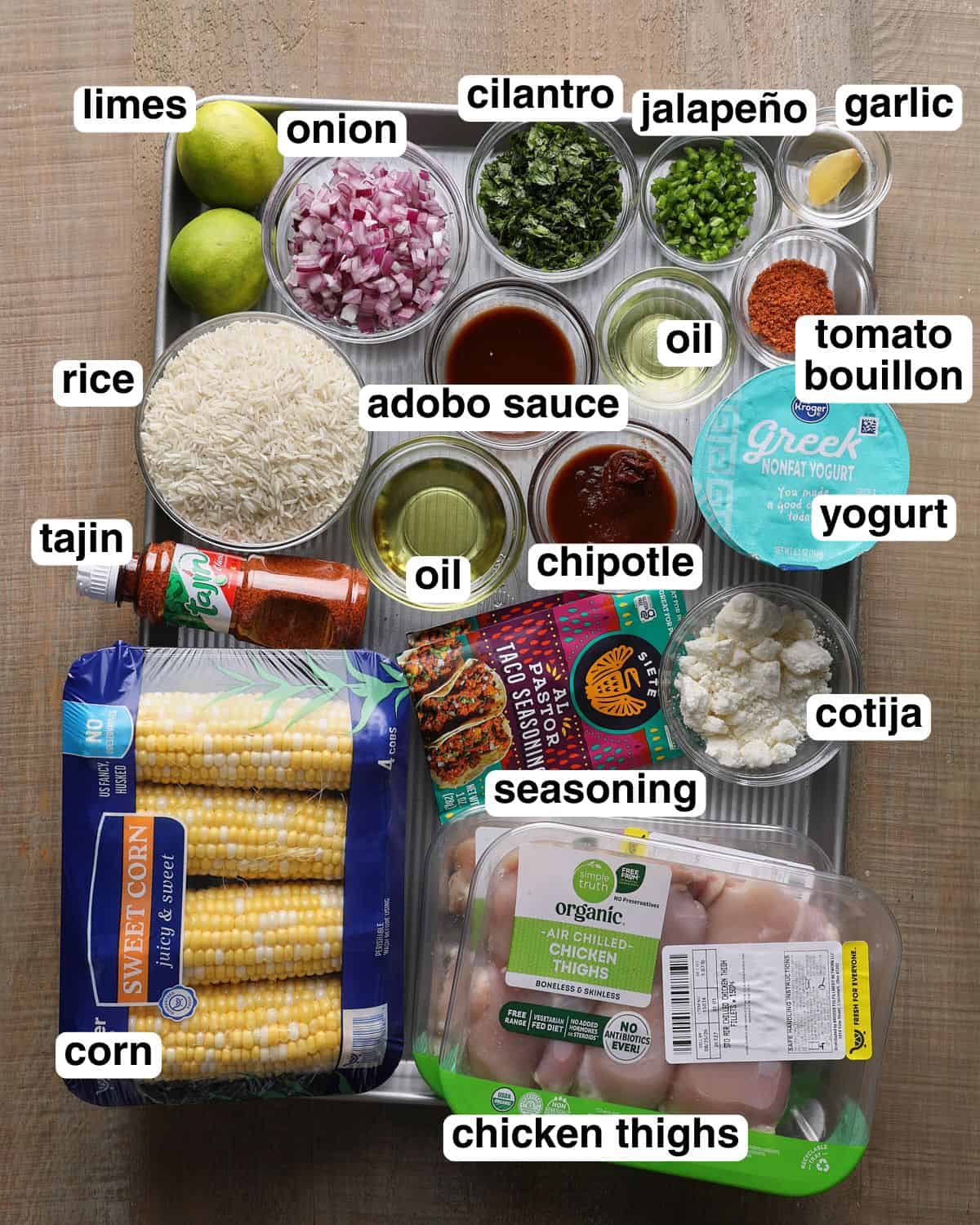 Labeled ingredients for street corn chicken rice bowls arranged on a sheet pan, including chicken thighs, corn, rice, yogurt, chipotle peppers, adobo sauce, seasoning, Cotija, Tajín, lime, cilantro, onion, jalapeño, garlic, tomato bouillon, and oil.