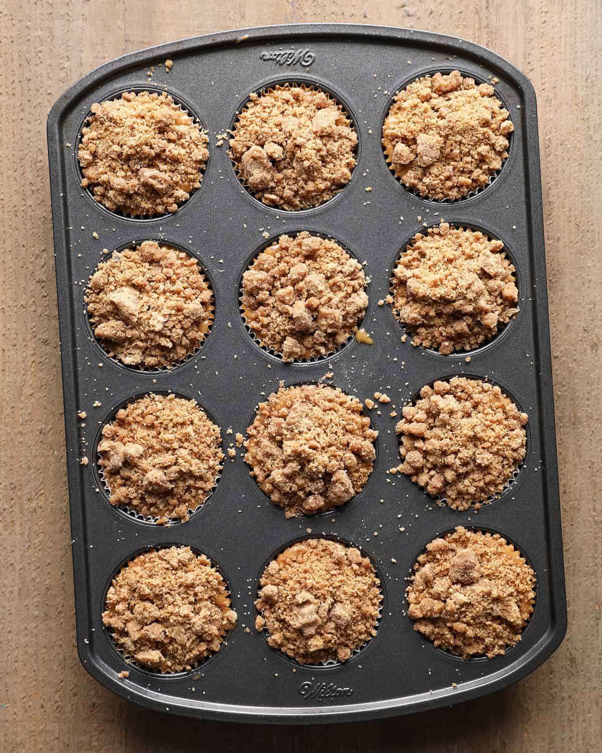 Twelve lined muffin wells filled with batter and topped with cinnamon crumb before baking.