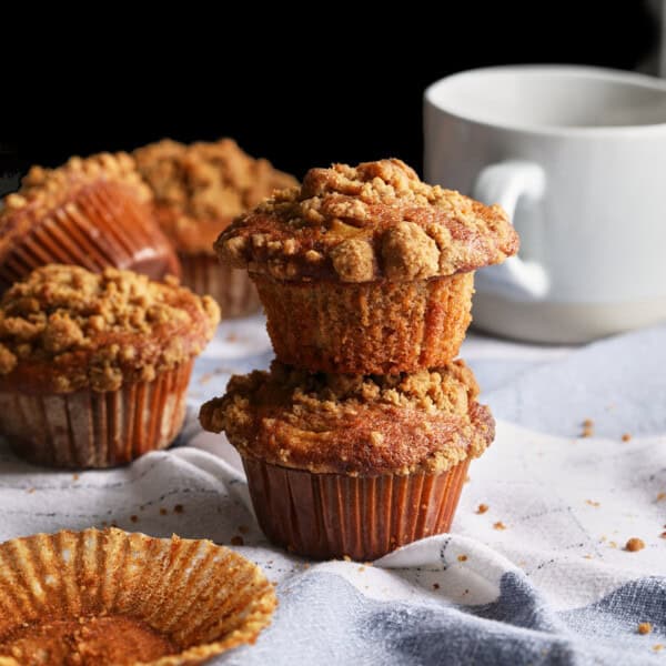 Apple cider muffins with tall cinnamon crumb tops stacked beside a mug.