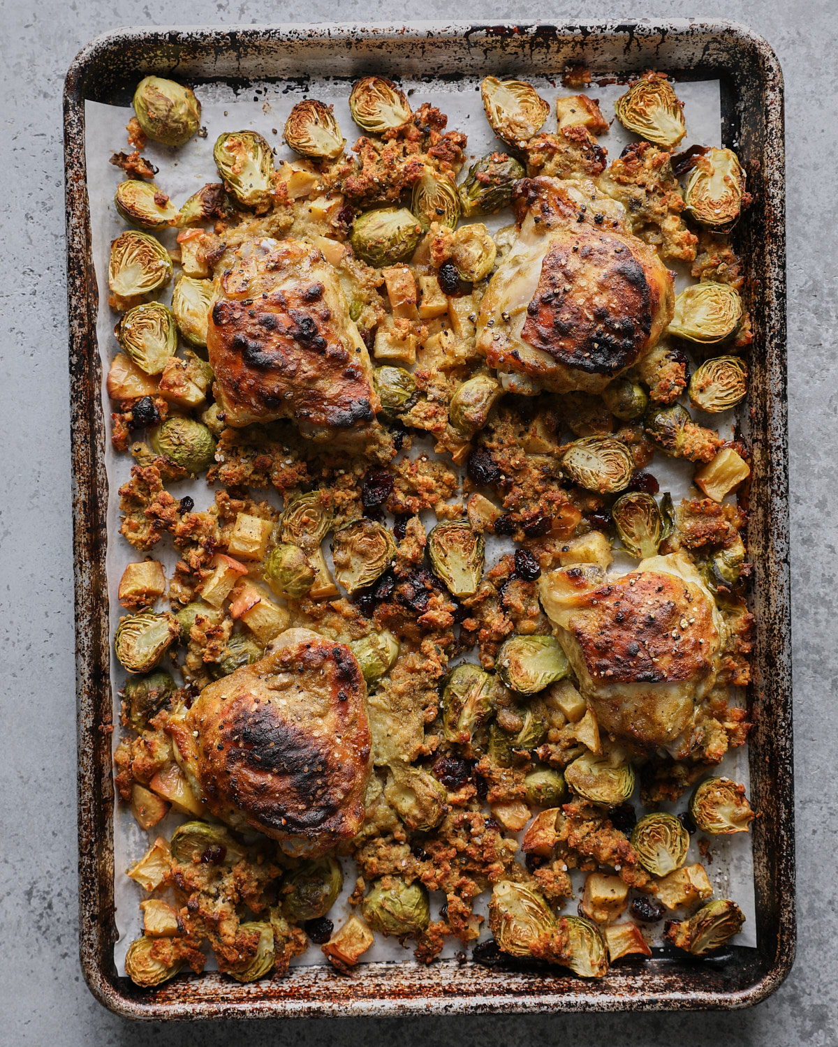 Browned chicken thighs over roasted stuffing, Brussels sprouts, apples, and cranberries on a parchment-lined sheet pan.