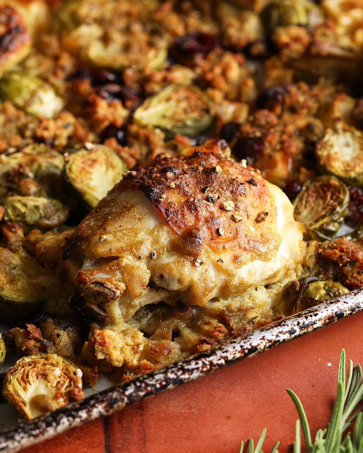 Browned chicken thigh resting above soft stuffing with crisp stuffing pieces and roasted Brussels sprouts around it.